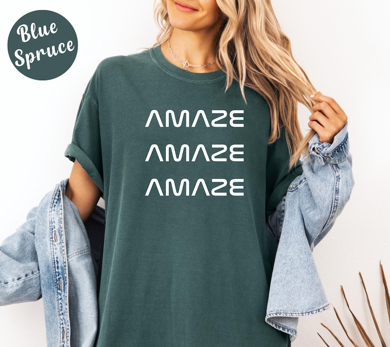 Pu&ograve; includere: Una maglietta color Blue Spruce con la parola "AMAZE" stampata tre volte in bianco. La maglietta ha una vestibilit&agrave; ampia ed &egrave; realizzata in un materiale morbido. La modella indossa una giacca di jeans.