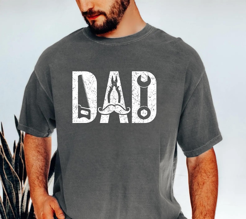 May include: Dark grey t-shirt featuring the word "DAD" in white. The letters are creatively designed with tools: a saw, pliers, and a wrench, with a moustache. A thoughtful gift for Father's Day.
