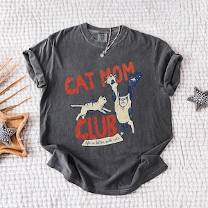 May include: Dark gray t-shirt with a distressed "Cat Mom Club" graphic in red and white. The design features two cats, one running and the other with its paws up. The text "Life is better with cats" is below the graphic.