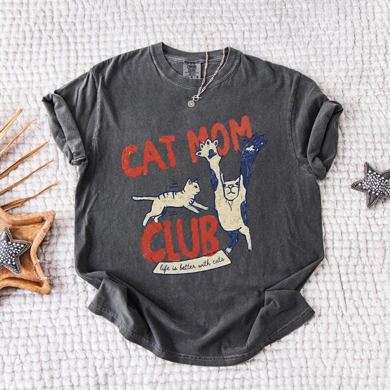 May include: Dark gray t-shirt with a distressed "Cat Mom Club" graphic in red and white. The design features two cats, one running and the other with its paws up. The text "Life is better with cats" is below the graphic.