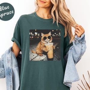 May include: Blue Spruce t-shirt featuring a graphic of a cat wearing sunglasses and drinking iced coffee. The cat is orange with black sunglasses. The shirt is a dark teal color.