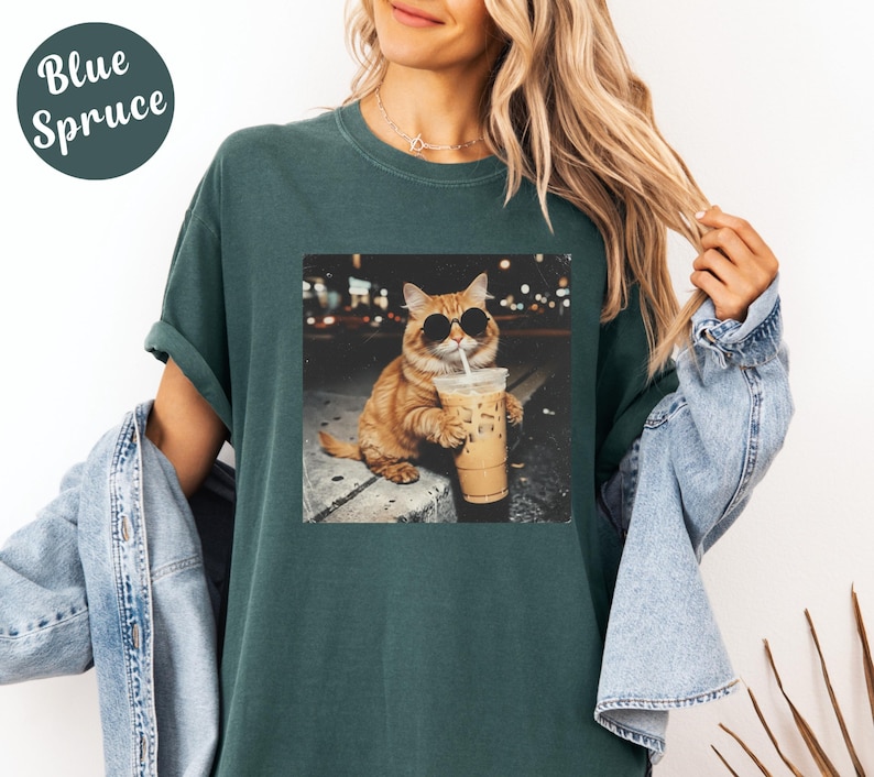 May include: Blue Spruce t-shirt featuring a graphic of a cat wearing sunglasses and drinking iced coffee. The cat is orange with black sunglasses. The shirt is a dark teal color.
