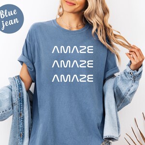 Pu&ograve; includere: Una maglietta blu jeans con la parola "AMAZE" stampata tre volte in bianco. La modella indossa una giacca di jeans sopra la maglietta. Un cerchio blu con le parole "Blue jean" si trova nell'angolo in alto a sinistra.