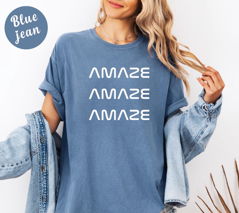 Pu&ograve; includere: Una maglietta blu jeans con la parola "AMAZE" stampata tre volte in bianco. La modella indossa una giacca di jeans sopra la maglietta. Un cerchio blu con le parole "Blue jean" si trova nell'angolo in alto a sinistra.
