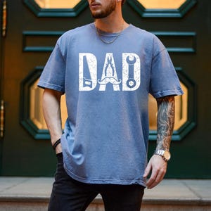 May include: A blue t-shirt with the word "DAD" in white, incorporating a saw, wrench, and other tools. The shirt has a distressed, vintage appearance.