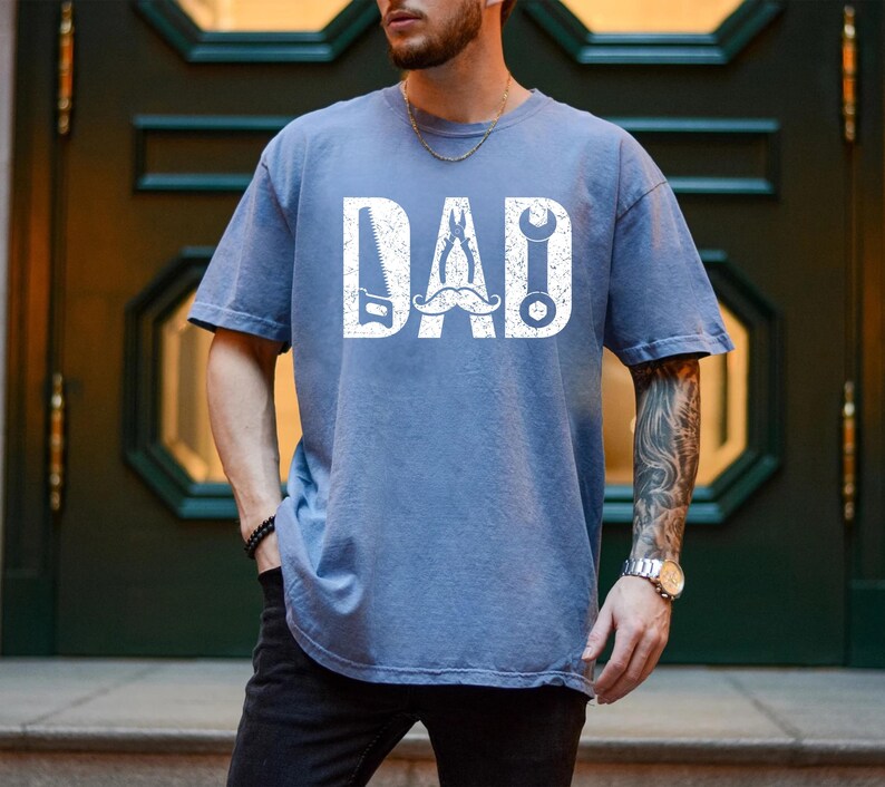May include: A blue t-shirt with the word "DAD" in white, incorporating a saw, wrench, and other tools. The shirt has a distressed, vintage appearance.