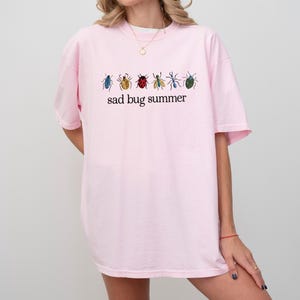 May include: A light pink t-shirt featuring a row of colorful insect illustrations above the text "sad bug summer" in black. The insects are in shades of blue, yellow, red, and green. The shirt is a casual, oversized fit.