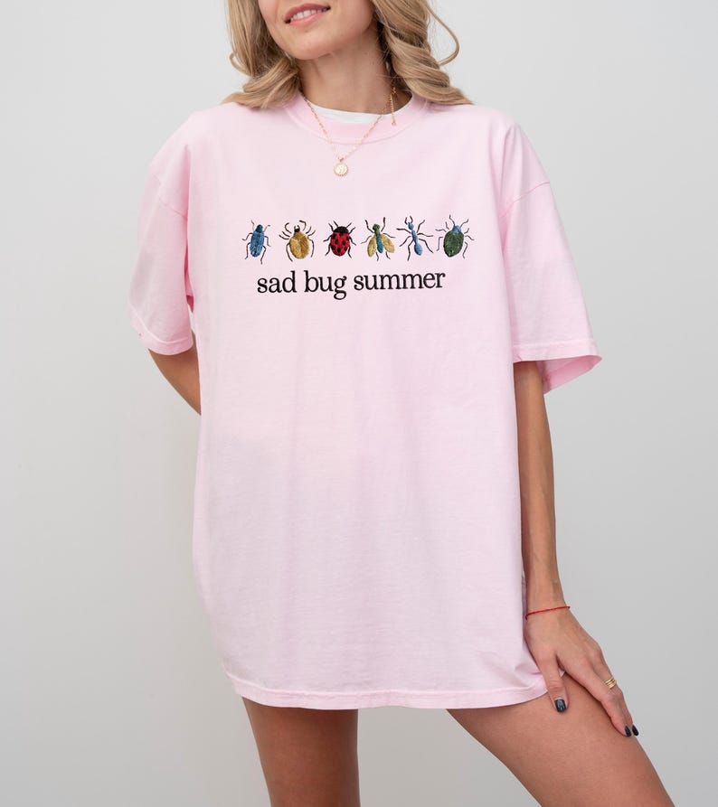 May include: A light pink t-shirt featuring a row of colorful insect illustrations above the text "sad bug summer" in black. The insects are in shades of blue, yellow, red, and green. The shirt is a casual, oversized fit.