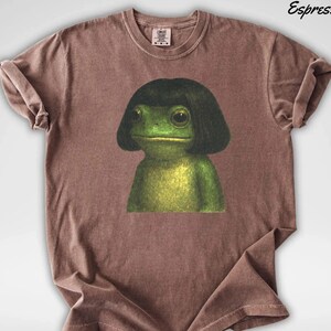 May include: Espresso-colored t-shirt featuring a whimsical illustration of a green frog with a black bob haircut. The frog has large, expressive eyes and is centered on the shirt. The shirt has a crew neck and short sleeves.