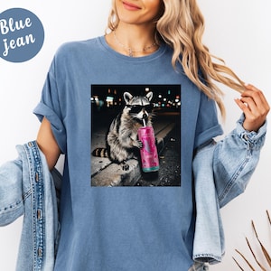May include: Blue jean t-shirt with a graphic of a raccoon wearing sunglasses and drinking from a pink can. The raccoon is sitting on a curb with a blurred city background. The shirt has a relaxed fit.
