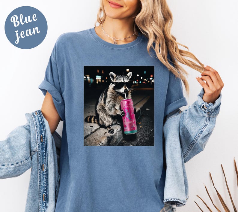 May include: Blue jean t-shirt with a graphic of a raccoon wearing sunglasses and drinking from a pink can. The raccoon is sitting on a curb with a blurred city background. The shirt has a relaxed fit.