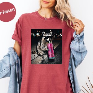 May include: Crimson-colored t-shirt featuring a graphic of a raccoon wearing sunglasses and drinking from a pink beverage container. The raccoon is sitting on a ledge with a city backdrop. The shirt is a casual style.