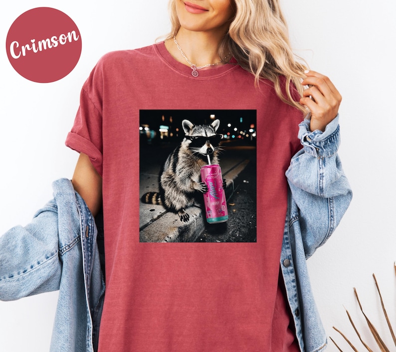May include: Crimson-colored t-shirt featuring a graphic of a raccoon wearing sunglasses and drinking from a pink beverage container. The raccoon is sitting on a ledge with a city backdrop. The shirt is a casual style.