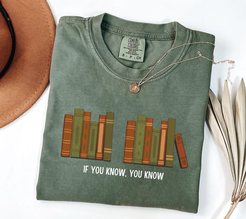 May include: Olive green t-shirt with a graphic of stacked books in shades of green and brown. The text "IF YOU KNOW, YOU KNOW" is printed below the books. A gold necklace with a pendant is draped across the shirt.