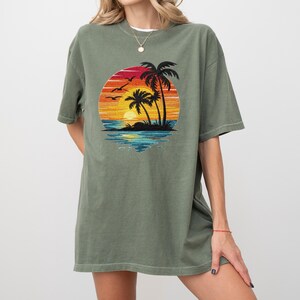 May include: Olive green t-shirt featuring a circular graphic of a sunset over the ocean with palm trees and birds. The graphic has vibrant colors of red, orange, yellow, and blue. The shirt has short sleeves and a relaxed fit.