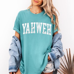 May include: Teal-colored t-shirt with the word "YAHWEH" printed in distressed white block letters. The shirt has short sleeves and a relaxed fit. A denim jacket is draped over the shoulders.
