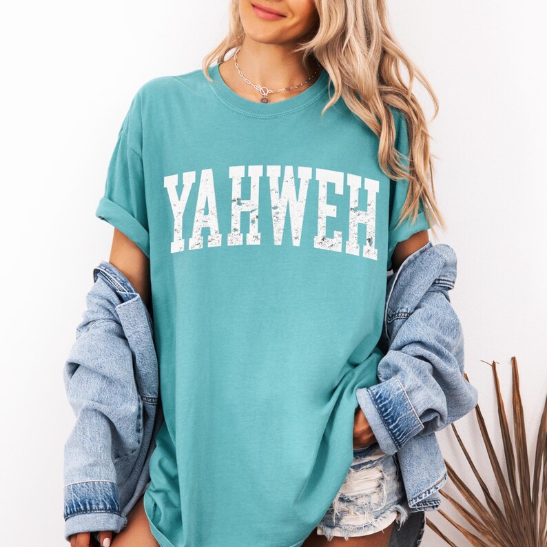May include: Teal-colored t-shirt with the word "YAHWEH" printed in distressed white block letters. The shirt has short sleeves and a relaxed fit. A denim jacket is draped over the shoulders.