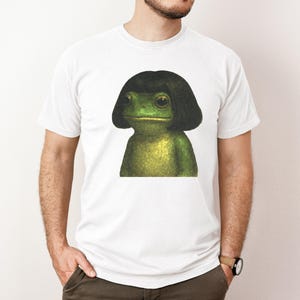 May include: White t-shirt featuring a green frog with a black wig. The frog has large eyes and a neutral expression. The shirt is a classic crew neck style.