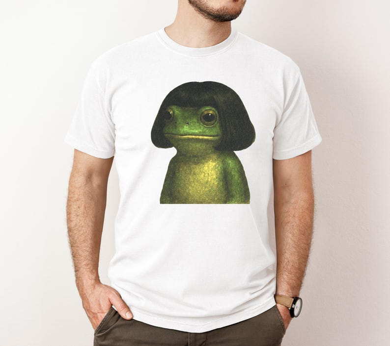 May include: White t-shirt featuring a green frog with a black wig. The frog has large eyes and a neutral expression. The shirt is a classic crew neck style.