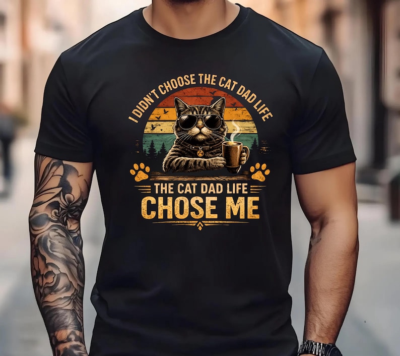 Op de afbeelding: Zwart t-shirt met een afbeelding van een kat met een zonnebril en een koffiemok. De tekst luidt: "I didn't choose the cat dad life, the cat dad life chose me."