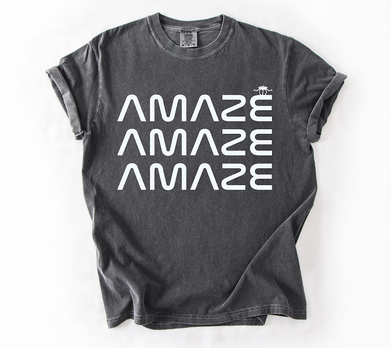 May include: Dark gray t-shirt with the word "AMAZE" stacked three times in white, with a small graphic above the last word. The shirt has rolled-up sleeves and a crew neck.