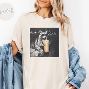May include: Ivory-colored t-shirt featuring a black and white graphic of a raccoon wearing sunglasses and drinking from a coffee cup. The raccoon is sitting on a ledge with a city backdrop. The shirt has a relaxed fit.