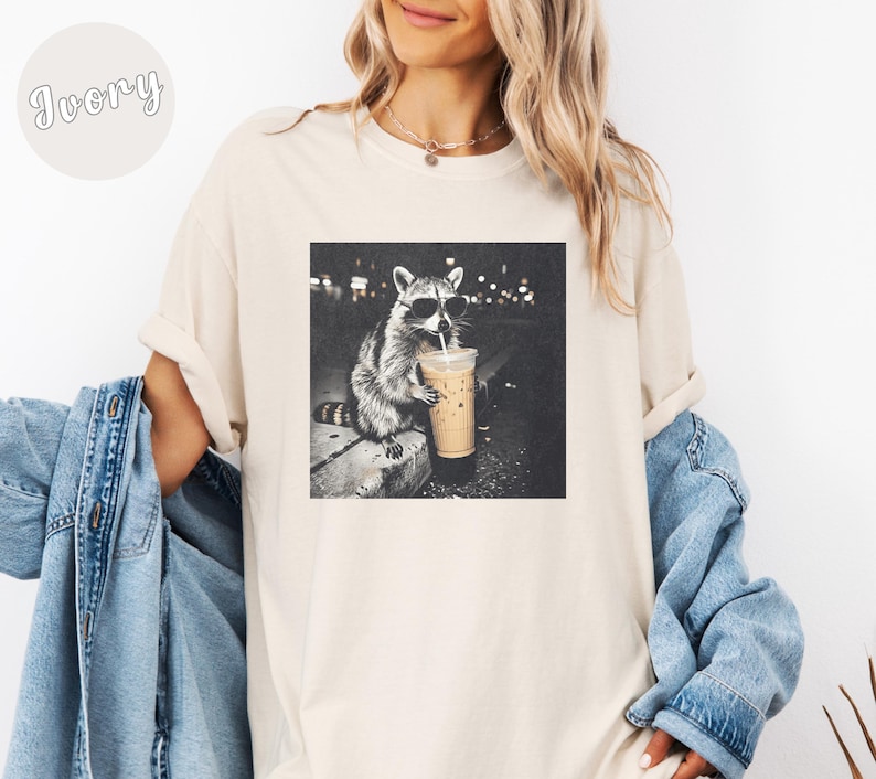 May include: Ivory-colored t-shirt featuring a black and white graphic of a raccoon wearing sunglasses and drinking from a coffee cup. The raccoon is sitting on a ledge with a city backdrop. The shirt has a relaxed fit.