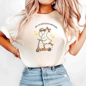 May include: Cream-colored t-shirt featuring a cartoon goose riding a pink scooter while drinking from a cup. The goose wears a pink bow and the text "OFF TO REGULATE EMOTIONS" is arched above the goose. Yellow starbursts are on either side.