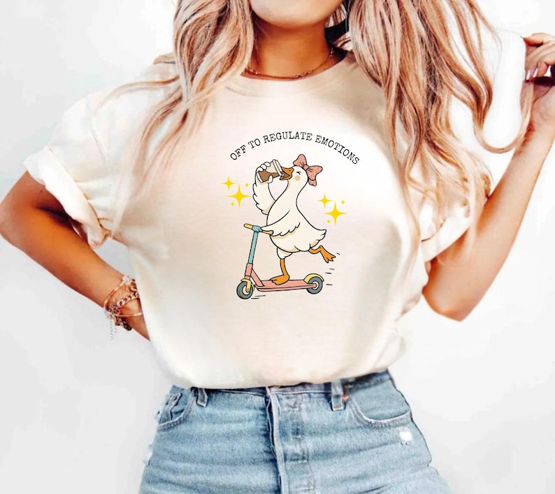 May include: Cream-colored t-shirt featuring a cartoon goose riding a pink scooter while drinking from a cup. The goose wears a pink bow and the text "OFF TO REGULATE EMOTIONS" is arched above the goose. Yellow starbursts are on either side.