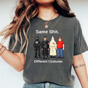 May include: Dark grey t-shirt featuring the text "Same Shit. Different Costume." and illustrations of four figures in various outfits. The figures include a person in tactical gear, a person in a black uniform, a person in a white robe, and a person in a red shirt.
