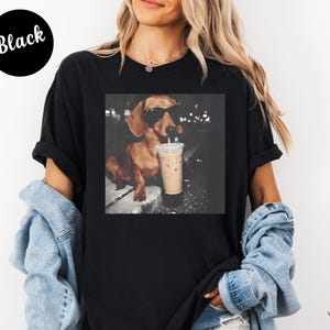 May include: Black t-shirt featuring a square image of a dachshund wearing sunglasses and holding a coffee drink. The dog is brown with black sunglasses. The text "Black" is in a black circle in the upper left corner.