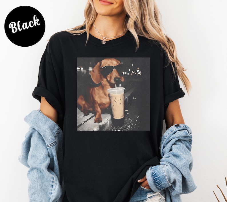 May include: Black t-shirt featuring a square image of a dachshund wearing sunglasses and holding a coffee drink. The dog is brown with black sunglasses. The text "Black" is in a black circle in the upper left corner.