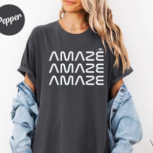 May include: Dark gray t-shirt with the word "AMAZE" stacked three times in white, with a small graphic above the top word. The shirt has rolled-up sleeves and is worn with a denim jacket.