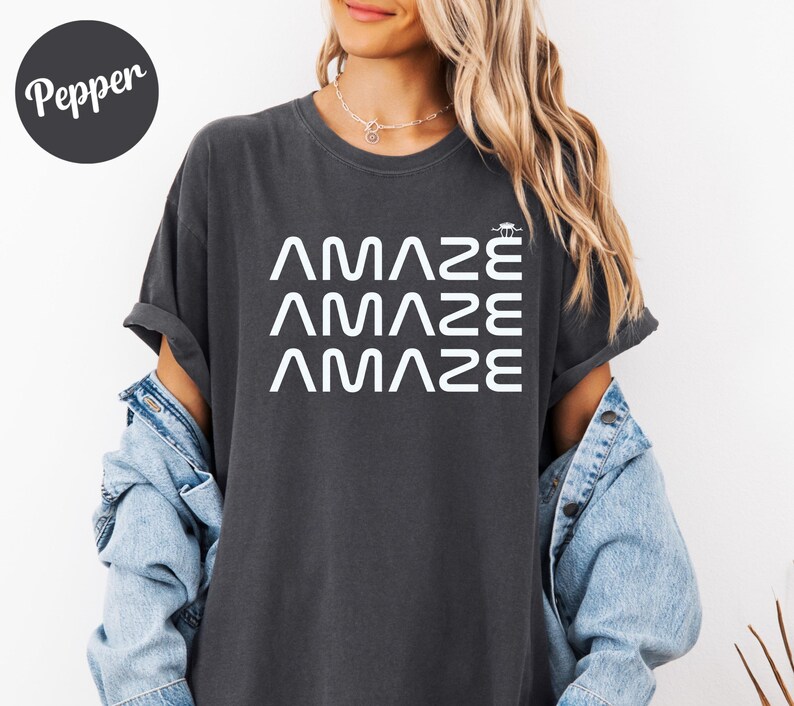 May include: Dark gray t-shirt with the word "AMAZE" stacked three times in white, with a small graphic above the top word. The shirt has rolled-up sleeves and is worn with a denim jacket.