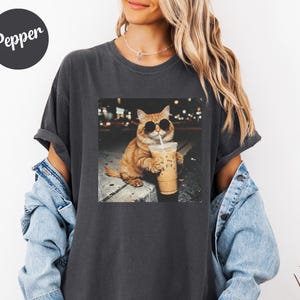 May include: Dark gray t-shirt featuring a graphic of an orange cat wearing sunglasses and drinking iced coffee. The image is set against a blurred city background. The word "Pepper" is printed on a black circle in the upper left corner.