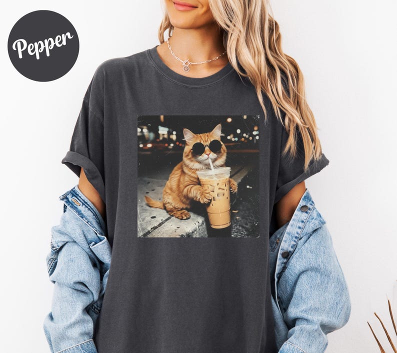 May include: Dark gray t-shirt featuring a graphic of an orange cat wearing sunglasses and drinking iced coffee. The image is set against a blurred city background. The word "Pepper" is printed on a black circle in the upper left corner.