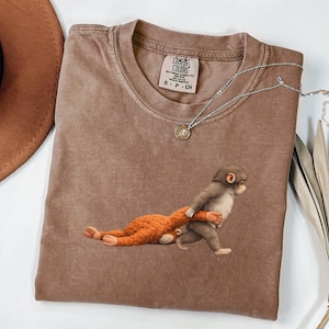 Puede incluir: Una camiseta marrón Comfort Colors con una ilustración caprichosa de dos monos. Un mono lleva al otro. Un collar plateado con un colgante está drapeado sobre la camisa. La camisa tiene una pequeña etiqueta que dice "Comfort Colors".