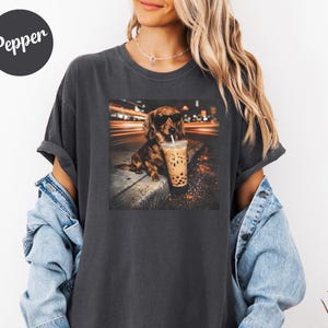 Puede incluir: Camiseta gris oscuro con un gr&aacute;fico de un perro salchicha con gafas de sol y sosteniendo un t&eacute; boba. El perro est&aacute; sentado en una acera con un fondo urbano borroso. La camiseta tiene la palabra "Pepper" en la parte superior izquierda.