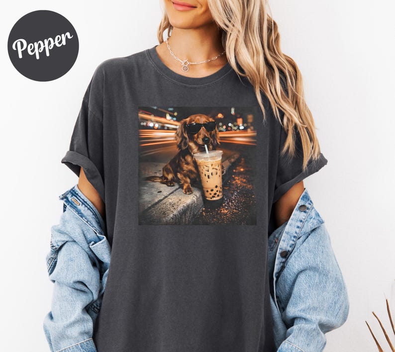 Puede incluir: Camiseta gris oscuro con un gr&aacute;fico de un perro salchicha con gafas de sol y sosteniendo un t&eacute; boba. El perro est&aacute; sentado en una acera con un fondo urbano borroso. La camiseta tiene la palabra "Pepper" en la parte superior izquierda.