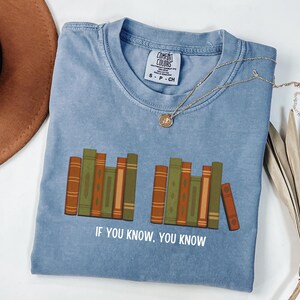 May include: A blue Comfort Colors t-shirt with a graphic of stacked books in shades of brown and green. The text "IF YOU KNOW, YOU KNOW" is printed below the books. A gold necklace with a pendant is on the shirt.