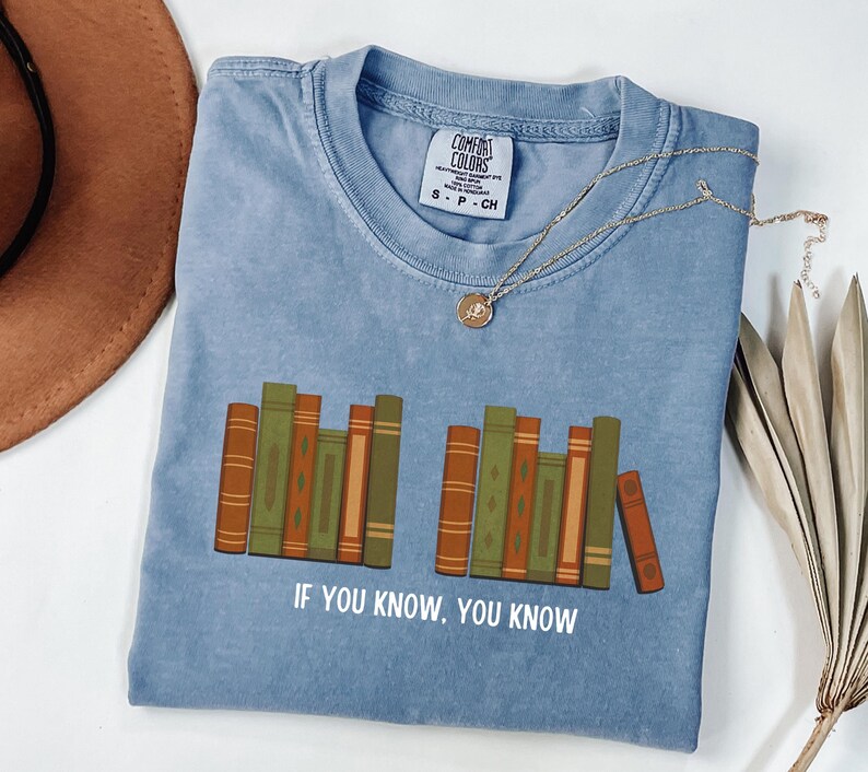 May include: A blue Comfort Colors t-shirt with a graphic of stacked books in shades of brown and green. The text "IF YOU KNOW, YOU KNOW" is printed below the books. A gold necklace with a pendant is on the shirt.