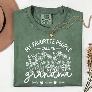 May include: A sage green t-shirt with the text "MY FAVORITE PEOPLE CALL ME grandma" in white script. The shirt features a floral design and the names "Emily", "Olivia", and "Noah". A gold star pendant necklace rests on the shirt.