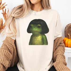 May include: Cream-colored t-shirt featuring a green frog with a black bob haircut. The frog has large eyes and a neutral expression. The shirt is a casual style, suitable for everyday wear.