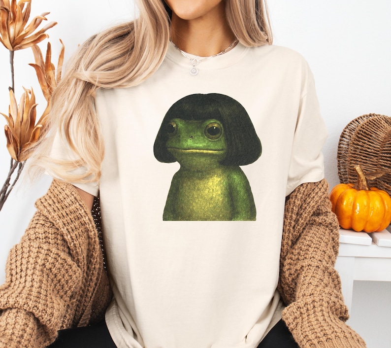 May include: Cream-colored t-shirt featuring a green frog with a black bob haircut. The frog has large eyes and a neutral expression. The shirt is a casual style, suitable for everyday wear.