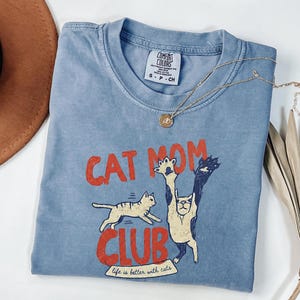 May include: A blue Comfort Colors t-shirt with the words "CAT MOM CLUB" in red, featuring cat illustrations. The shirt also has the phrase "life is better with cats." A gold necklace with a pendant is on the shirt.