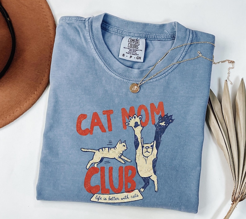 May include: A blue Comfort Colors t-shirt with the words "CAT MOM CLUB" in red, featuring cat illustrations. The shirt also has the phrase "life is better with cats." A gold necklace with a pendant is on the shirt.