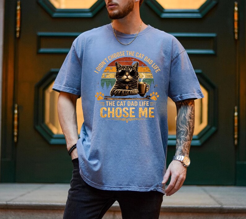 Op de afbeelding: Blauw T-shirt met een afbeelding van een kat met een zonnebril en een koffiekopje. De tekst op het shirt luidt: "Ik heb niet voor het kattenvaderleven gekozen, het kattenvaderleven koos mij."