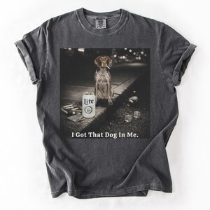 Pu&ograve; includere: T-shirt grigio scuro con una grafica di un cane con una sigaretta, una lattina di birra Lite e la scritta "I Got That Dog In Me." L'immagine &egrave; stampata su una maglietta morbida e confortevole.