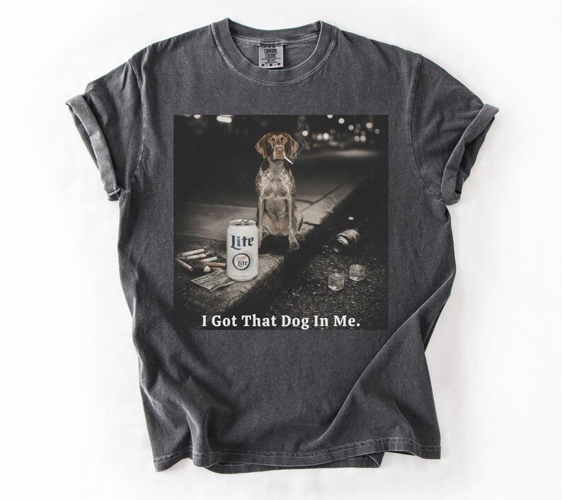 Pu&ograve; includere: T-shirt grigio scuro con una grafica di un cane con una sigaretta, una lattina di birra Lite e la scritta "I Got That Dog In Me." L'immagine &egrave; stampata su una maglietta morbida e confortevole.