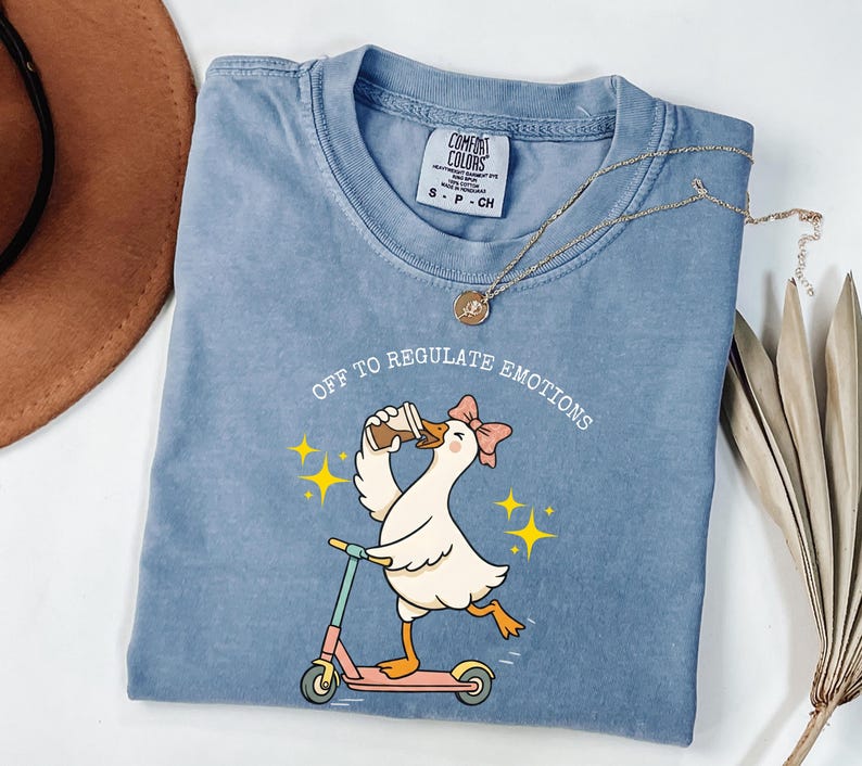 May include: A blue Comfort Colors t-shirt featuring a cartoon goose riding a scooter and drinking coffee. The text "OFF TO REGULATE EMOTIONS" is arched above the goose. A gold necklace is draped across the shirt.